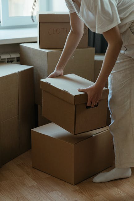 A person wearing light-colored clothing is seen lifting a small cardboard box in an indoor space with natural light coming through a window. The boxes are plain brown with no labels or markings, except for one that has the word 'CLOTHES' written on it in red ink. The boxes are stacked on top of each other, with some positioned on a wooden floor and others on a larger cardboard base. The individual appears to be involved in packing or preparing for a home relocation, indicative of furniture transport and packing activities typical in house removals. Visible in the background are additional cardboard boxes and soft furnishings, such as a fabric-covered piece of furniture, suggesting an ongoing move or packing process. Man and a Van Barnet regularly support clients with efficient removals near High Barnet station, using appropriate packing and loading procedures during furniture transport and home moving logistics.