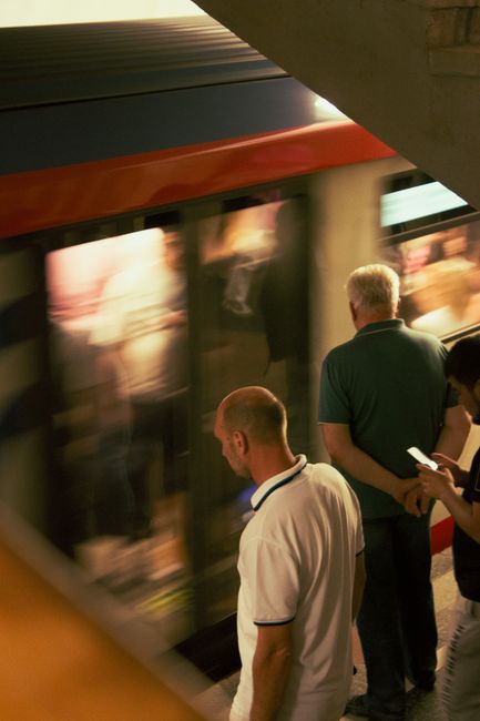 The image shows three individuals waiting on a platform next to a approaching train, which is passing by at high speed, causing a motion blur effect on the train’s exterior and the reflections in the windows. One person, a man with short hair, is wearing a navy polo shirt and is holding a smartphone, while another man with a bald head is dressed in a light-colored polo shirt and is looking at the train as it arrives. A woman with dark hair is partially visible, also facing the train. The platform appears to be indoors with artificial lighting, and the scene captures a moment of home relocation or moving logistics, implying a journey for furniture or personal belongings. No specific furniture or packaging materials are visible in this particular image, but the passing train provides context for transit associated with removals and moving services. Occasionally, Man and a Van Barnet's services relate to such transport and logistics for house moves near High Barnet station.