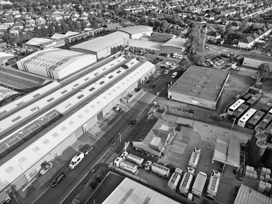 An aerial view in black and white showing a commercial area with multiple large industrial warehouses and smaller adjacent buildings. Several parked vehicles, including vans, cars, and trucks, are vis