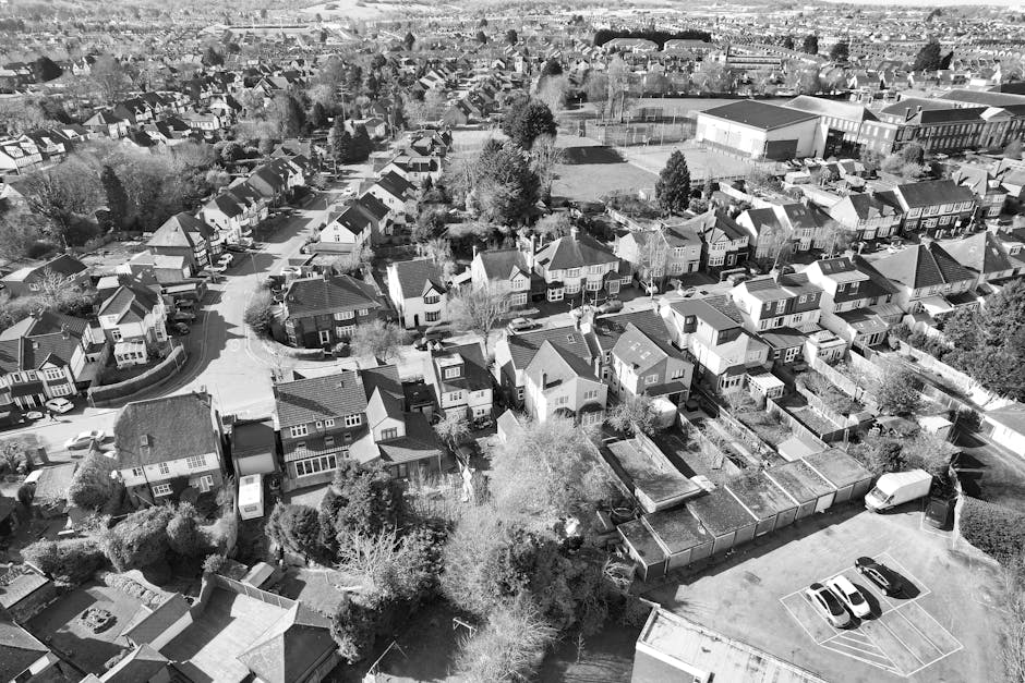 An aerial view in black and white showing a commercial area with multiple large industrial warehouses and smaller adjacent buildings. Several parked vehicles, including vans, cars, and trucks, are vis