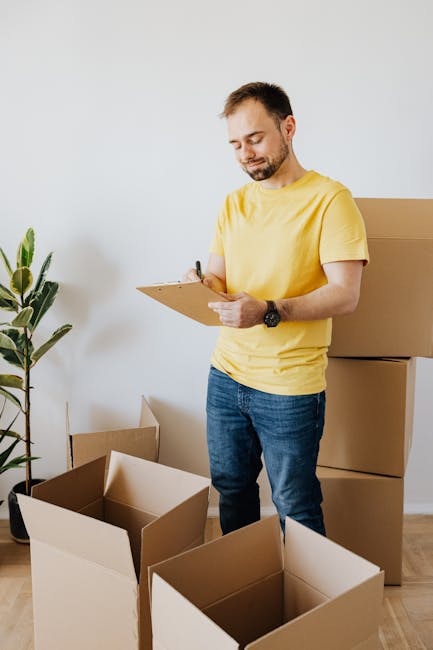A man with short dark hair and a beard, wearing a yellow T-shirt and blue jeans, is standing indoors next to several open cardboard boxes and a few sealed boxes stacked behind him. He is holding a cli